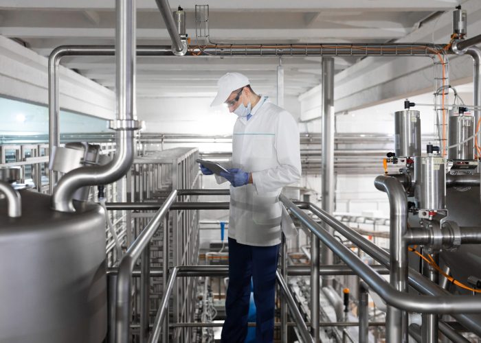 Technologist in a white robe, gloves and mask holding a tablet and stands near the bedon factory dairy products. The inspector checks the performance at the production plant