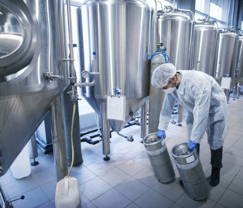 Worker in protective suit carrying chemicals in metal reservoirs.